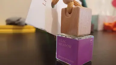Small glass Lilac Meadow car fragrance bottle with a purple label and wooden cap, on a dark surface.