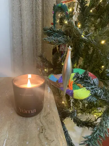 A lit YUMA Black Orchid scented candle in a black jar beside a decorated Christmas tree.