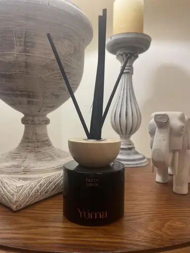 YUMA Fresh Linen black room diffuser with a wooden lid and black reeds on a wooden surface.