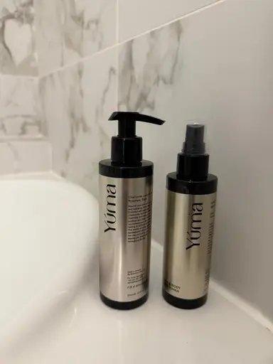 Two YUMA metallic bottles with black pumps placed beside a white sink and marble wall.