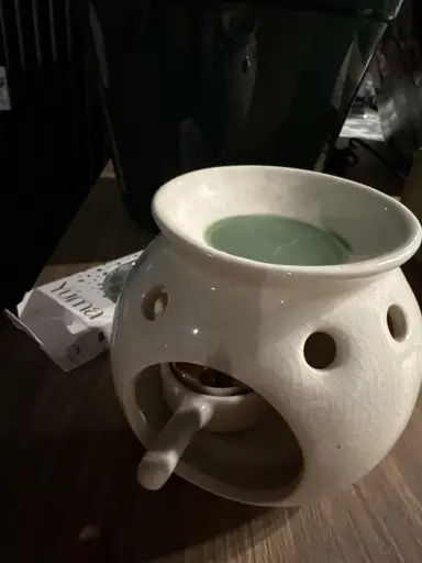 A white ceramic burner with melted green wax on top and a lit tea light candle inside.