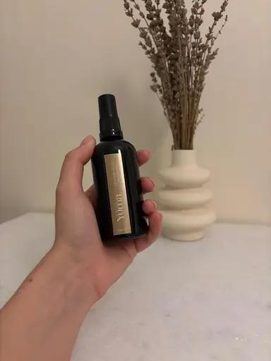 A hand holds the YUMA Eternal Eden room spray in front of a vase with dried lavender.