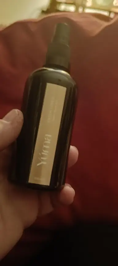 A hand holding a dark YUMA room spray bottle with a gold label.