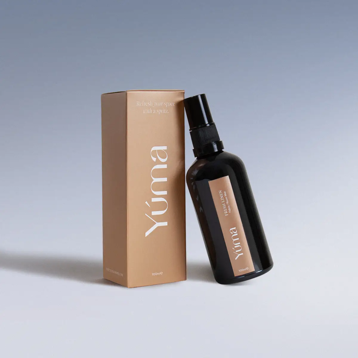 Dark glass bottle of YUMA Fresh Linen Fabric/Room Mist beside its beige paper packaging on a light blue background.
