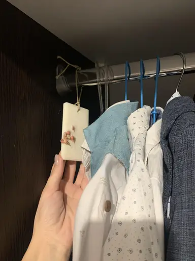A white scented wardrobe sachet with dried flowers hanging beside shirts.