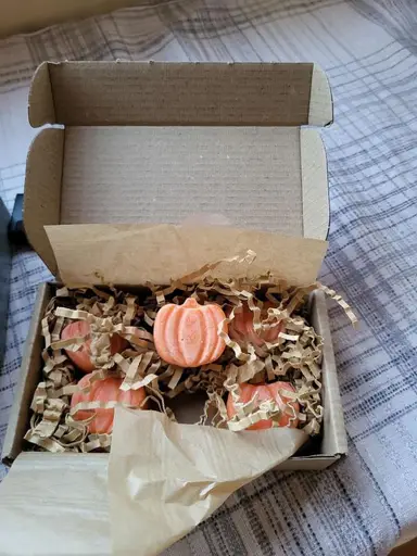 An open paper box with orange pumpkin-shaped wax melts resting on brown paper packaging material.