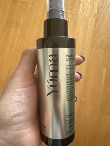 A hand holding a cylindrical YUMA Hair & Body Mist bottle with a silver label.