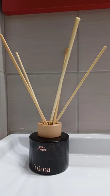 A black glass YUMA reed diffuser with a wooden cap and five reeds, set against grey tiles.