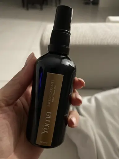 A hand holding the YUMA Salted Caramel Room Mist in a black glass bottle with a gold label.