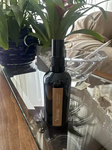 A black YUMA room spray bottle on a glass table next to indoor green plants.