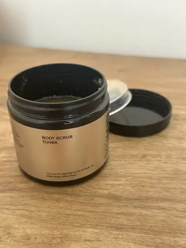 An open jar of YUMA's Body Scrub Tonka with a gold label, resting on a wooden surface.