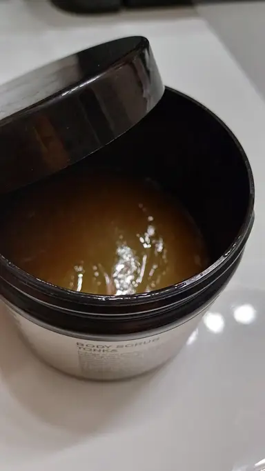 Open dark jar containing a brown body scrub with a grainy texture, on a white background.