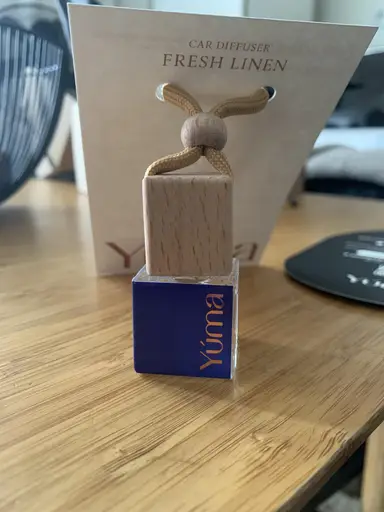 Small YUMA glass car air freshener with a wooden cap and cord, placed on a wooden surface.