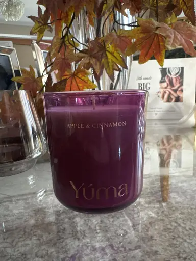 YUMA Apple & Cinnamon scented candle in a purple glass jar, placed in front of autumn leaves.
