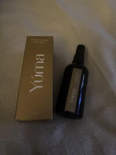 A dark glass Eternal Eden Room Mist bottle beside its gold packaging, resting on fabric.