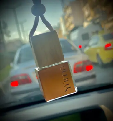 A YUMA hanging car fragrance with a wooden cap and glass bottle, set against a blurred background of moving cars.