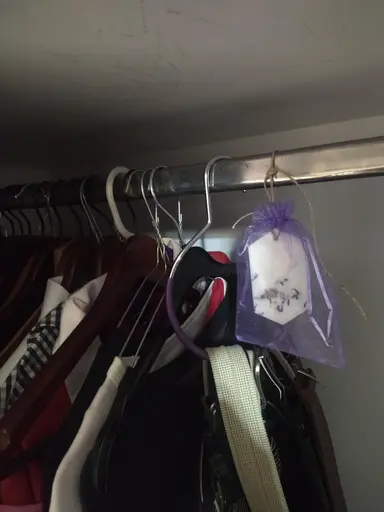 A white scented wardrobe sachet with dried flowers inside a purple pouch, hanging on a rod with hangers.