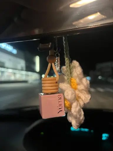 Pink YUMA car diffuser hanging from the mirror next to a woven white daisy.