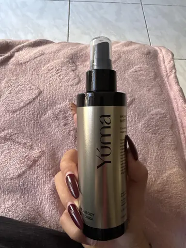 A hand holding a YUMA metallic spray bottle above a pink towel.