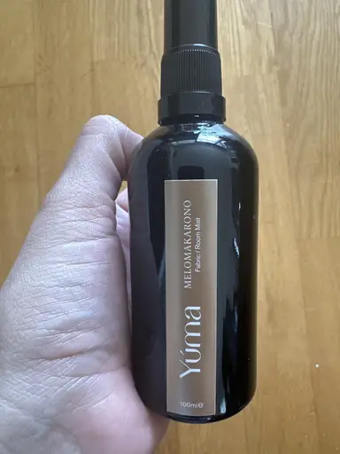 A hand holding the YUMA Melomakarono Room Mist black bottle in front of a wooden floor.