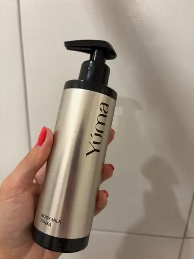 A hand holding YUMA's Body Milk Tonka in a metallic bottle with a black pump.