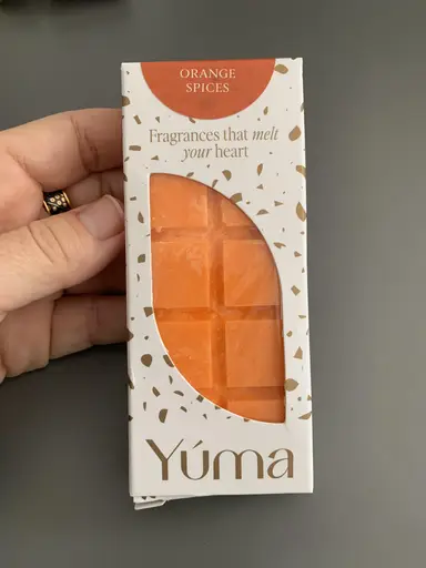 A hand holding the white YUMA Orange Spices packaging with orange wax melts.
