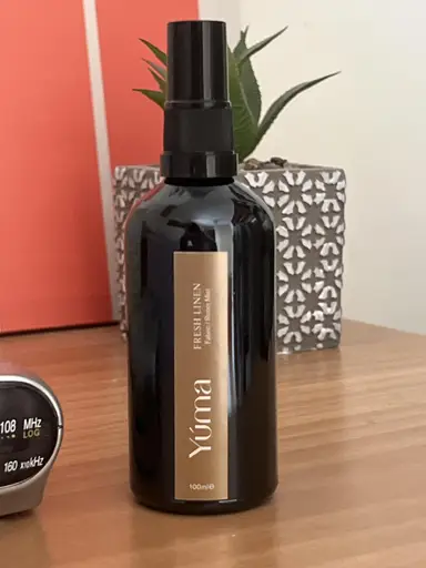A black Yuma Fresh Linen room spray bottle on a wooden surface in front of a decorative plant.