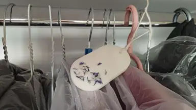 Oval white wardrobe sachet with dried blue petals, hanging by twine between coat hangers.