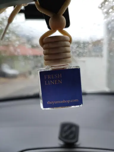 Fresh Linen car air freshener with a wooden cap hanging on a rearview mirror in front of a rain-streaked window.