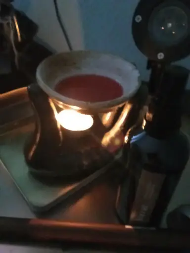 Ceramic wax melt burner with a lit tealight and red liquid, next to a dark pump bottle.