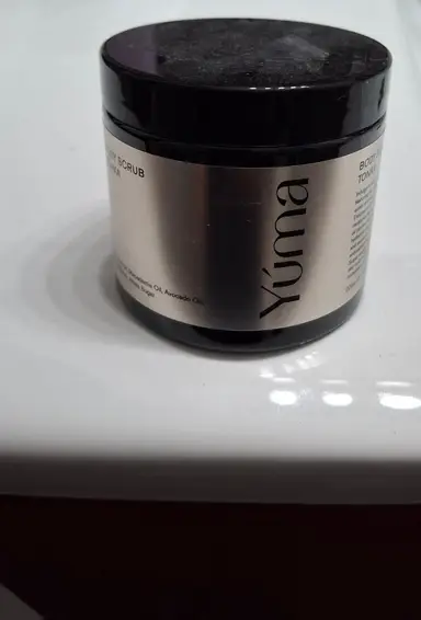 A black YUMA body scrub jar with a silver label, placed on a white surface.
