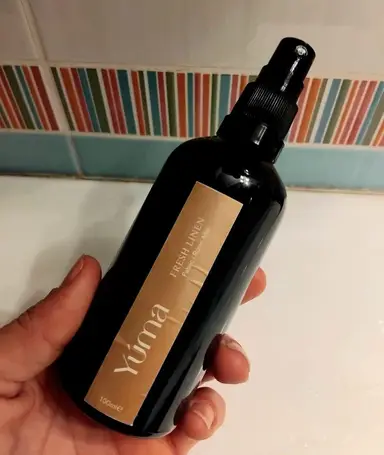 A hand holding the YUMA Fresh Linen room spray in a dark bottle with a beige label.
