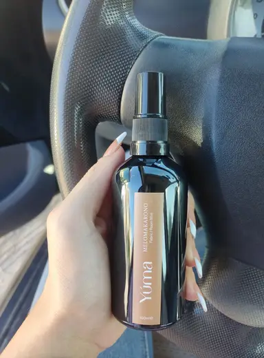 A woman's hand holding YUMA Melomakarono Room Mist in front of a car steering wheel.