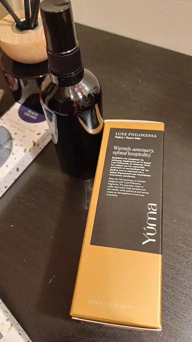 A black spray bottle next to the gold and black YUMA Luxe Philoxenia packaging on a dark surface.
