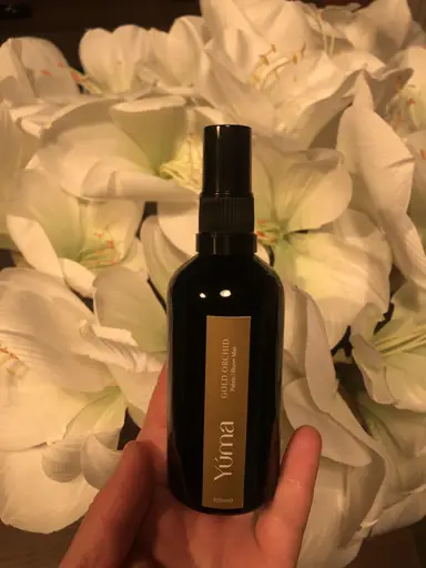 A hand holds the YUMA Gold Orchid room mist in front of white lily blossoms.