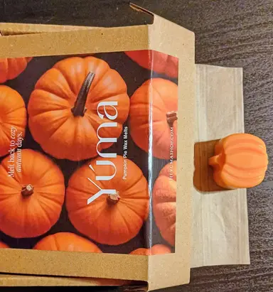 A small orange pumpkin-shaped candle next to a YUMA paper box featuring pumpkin imagery.