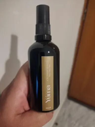 A hand holding the YUMA Cycladic Roots Room Mist in a black bottle with a gold label.