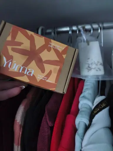 A hand holding a YUMA Gold Orchid wardrobe sachet box in front of hanging clothes.