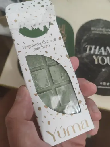 A hand holding a white YUMA box containing green chocolate-shaped wax melts.