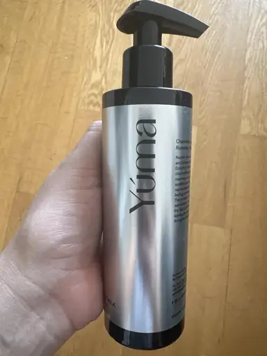 A hand holding a silver YUMA bottle with a black pump over a wooden floor.