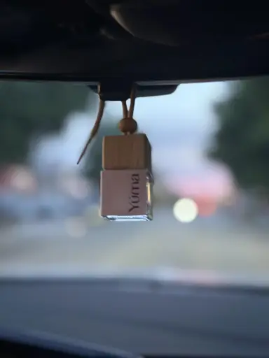 A small YUMA glass car fragrance with a wooden cap hanging from a rearview mirror.