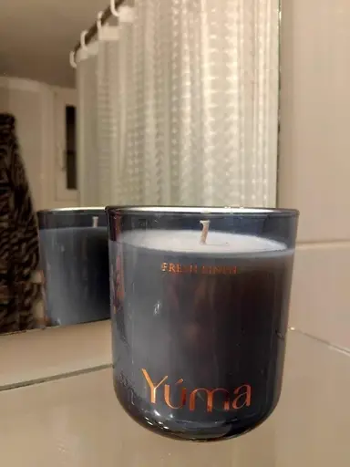 YUMA Fresh Linen scented candle in a dark glass vessel in front of a bathroom mirror.