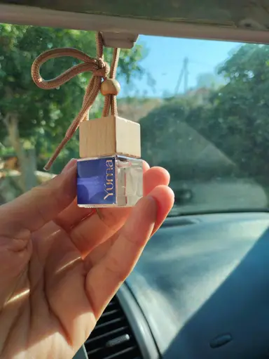 A hand holding a small YUMA glass car diffuser with a wooden stopper and brown cord.