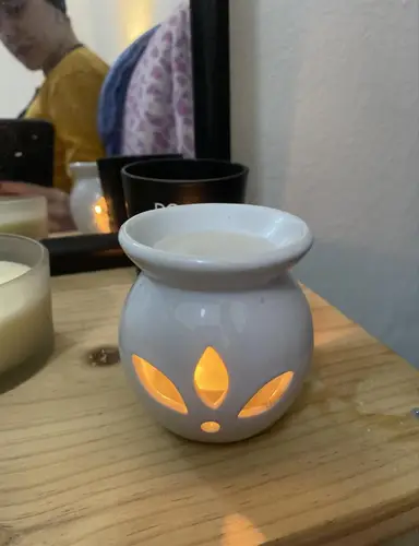 White ceramic oil burner with a lit tealight on a wooden surface next to candles.