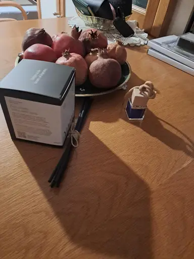 A black room fragrance box next to black diffuser sticks and a bowl of pomegranates.