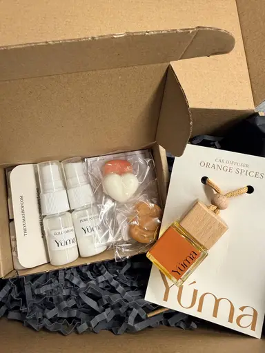 An open YUMA gift box containing fragrance sprays, heart-shaped wax melts, and an Orange Spices car air freshener.