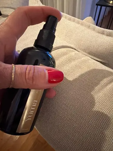 A hand with red nail polish holding a YUMA black room spray bottle above a fabric sofa.