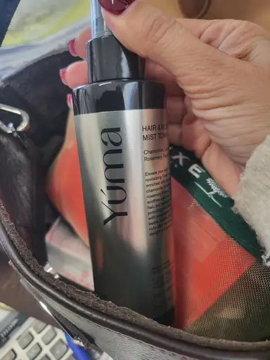 A woman's hand holding a silver YUMA Hair & Body Mist bottle inside a leather bag.