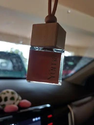 A small YUMA glass car fragrance bottle with a wooden cap hanging from a mirror.