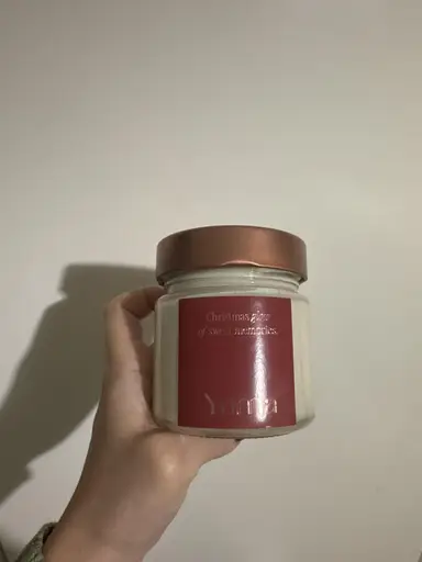 A hand holding a small YUMA glass jar with a red label and copper lid in front of a white wall.
